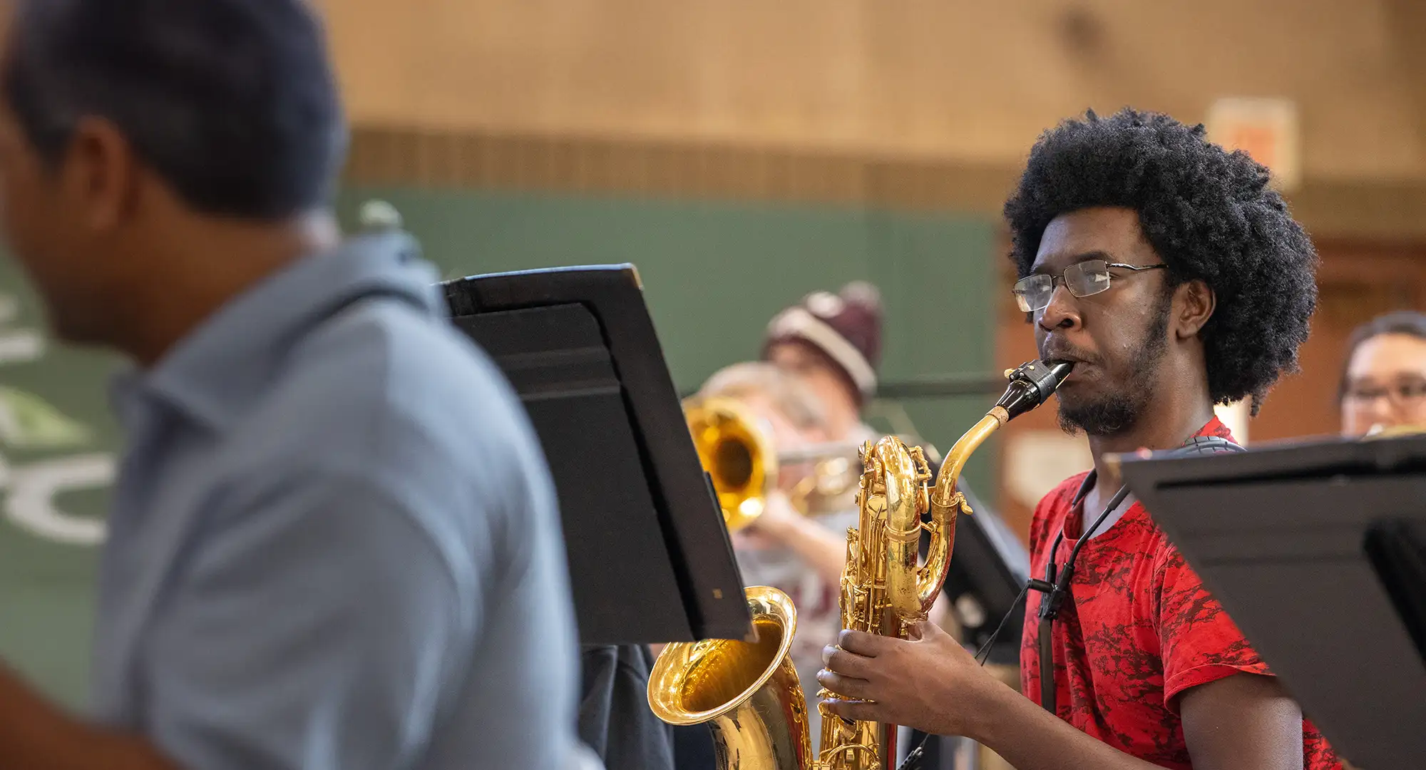 Student playing a saxophone in a campus wind ensemble rehearsal, with music stands and other musicians in the background.
